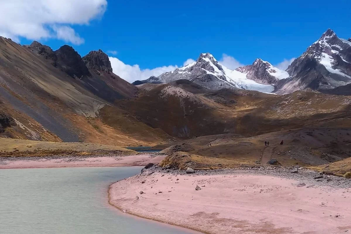 The stunning beauty of the high altitude Andes on the Ausangate route A breathtaking panoramic landscape of the Peruvian Andes during the Ausangate trek. It features a turquoise glacier lake, high altitude puna grasslands, and several snow-capped mountain ranges in the distance.