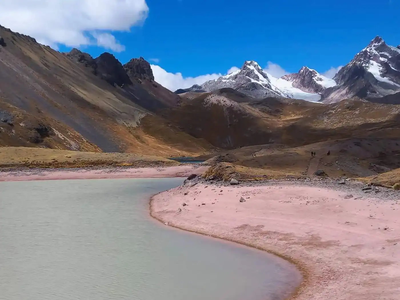 The stunning beauty of the high altitude Andes on the Ausangate route A breathtaking panoramic landscape of the Peruvian Andes during the Ausangate trek. It features a turquoise glacier lake, high altitude puna grasslands, and several snow-capped mountain ranges in the distance.