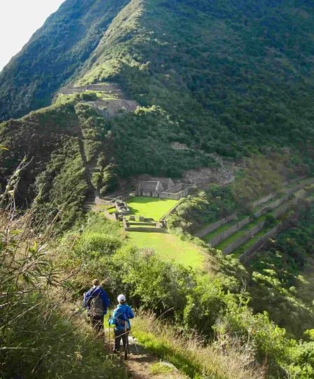 The remote and magnificent 'cradle of gold', Choquequirao A magnificent long-distance panoramic overview of the remote Choquequirao archaeological park. Extensive stone terraces cling to a steep Andean mountain ridge overlooking a deep green valley and distant mountains under a cloudy sky. A small group of travelers is visible, highlighting its lack of crowds.